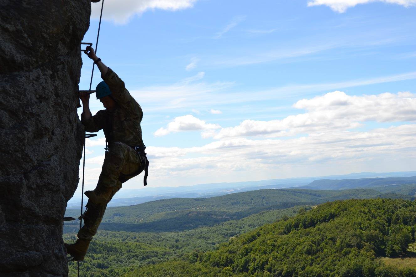 A climber in uniform ascends a rock face via ferrata above forested hills.